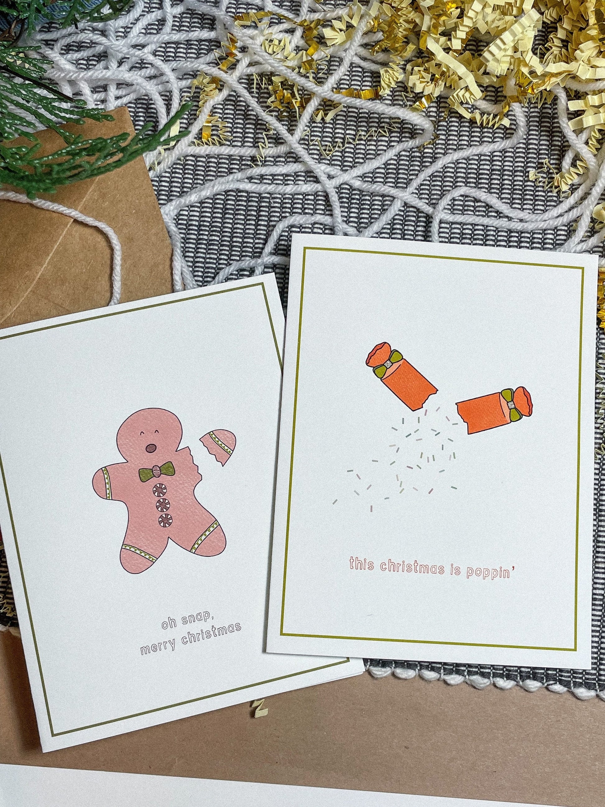 white greeting card with a green border and a little pink gingerbread man with his arm snapping off and the text saying "oh snap, merry christmas - also showing the redish orange party popper card - a party popper mid pop with confetti spilling out