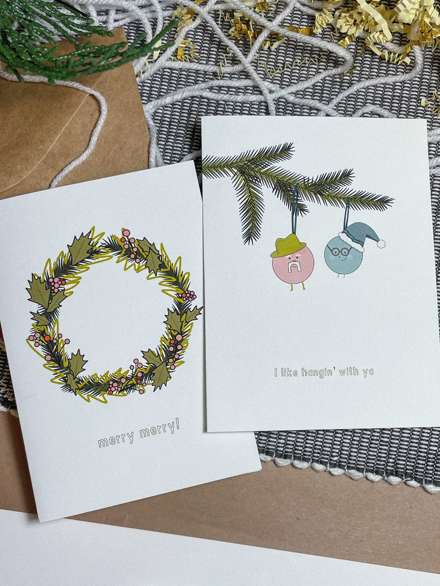 This card features a very merry Christmas wreath with the text 'merry merry!" It's the perfect Christmas card for anyone in your life! Shown alongside another card from the collection which shows 2 little ornament men hanging from a christmas tree branch