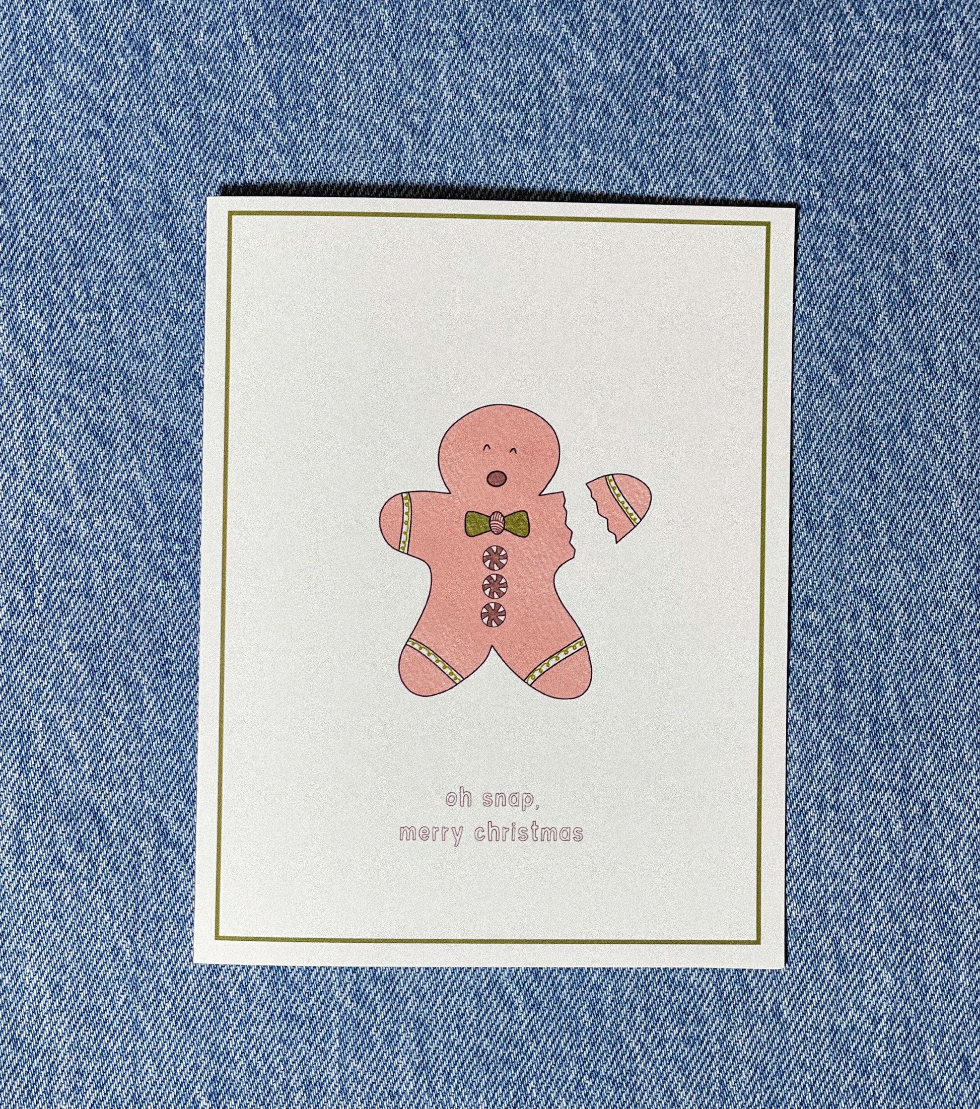 white greeting card with a green border and a little pink gingerbread man with his arm snapping off and the text saying "oh snap, merry christmas