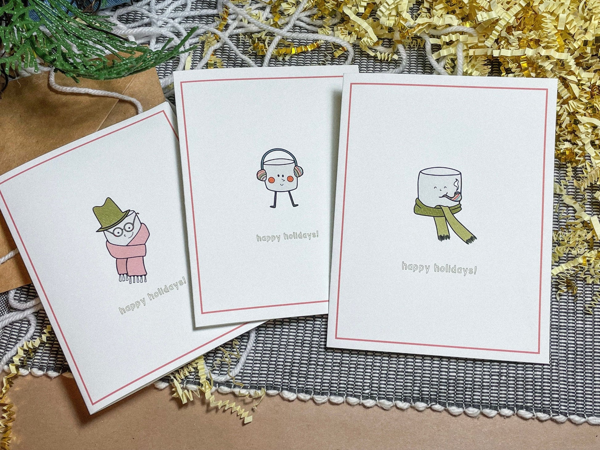 This holiday card features a drawing of a cute little marshmallow man with a scarf and a top hat with the text 'happy holidays!" Here you can see all 3 of the marshmallow cards - one with a hat and a scarf, one with little earmuffs, and one with a scarf and a pipe