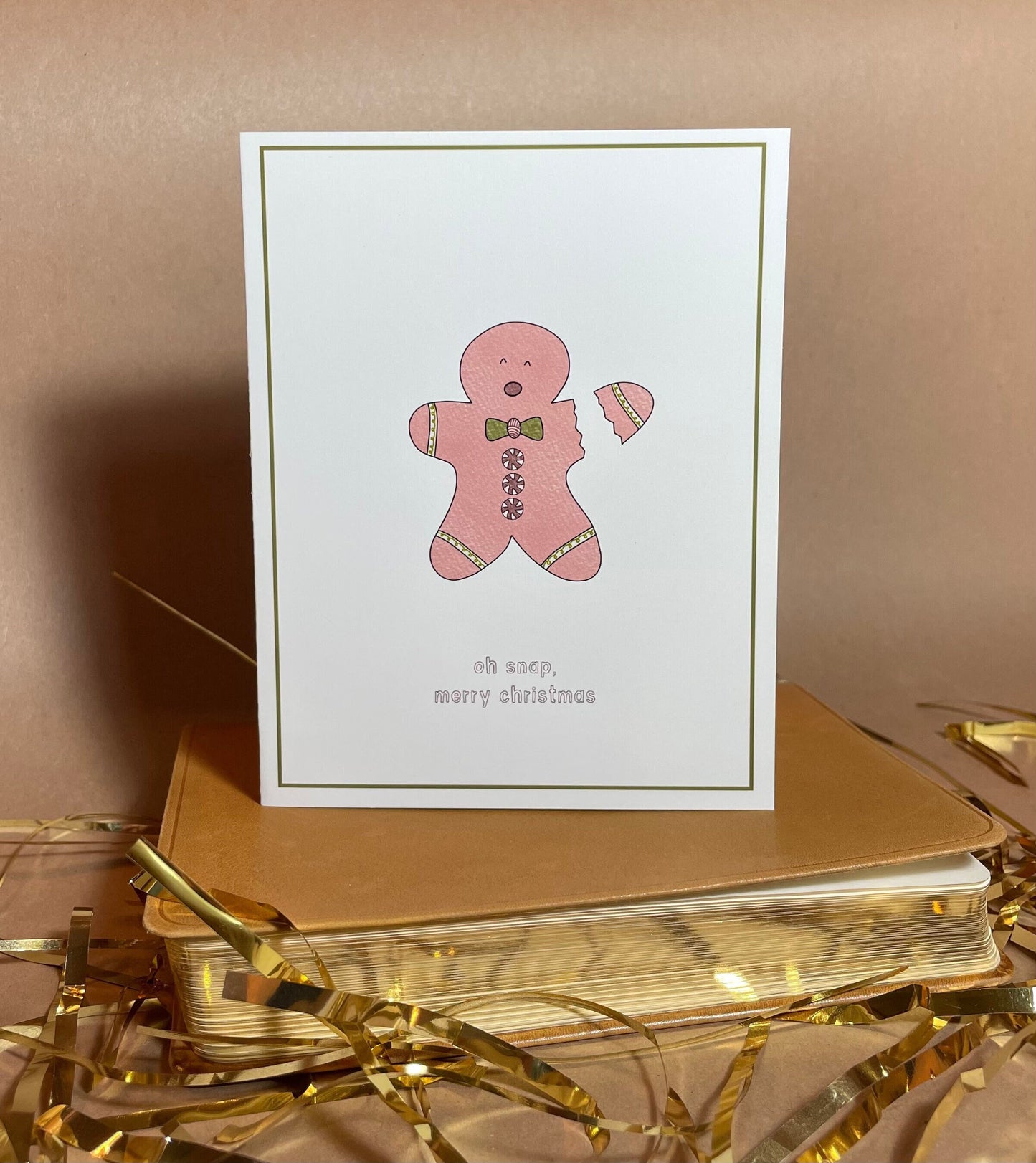 white greeting card with a green border and a little pink gingerbread man with his arm snapping off and the text saying "oh snap, merry christmas